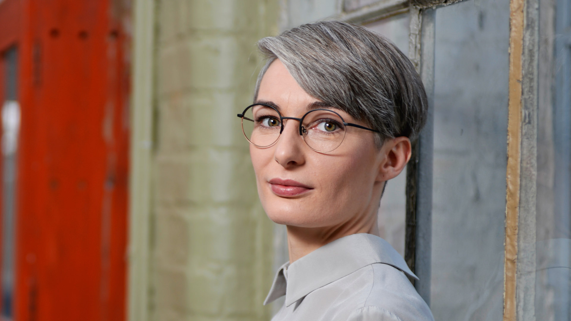 Junge Frau mit grauem Haar und stylischer runden Brille lehnt an eine Mauer