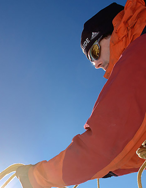Mann mit Sportbrille und Kletterausrüsting vor blauem Himmel